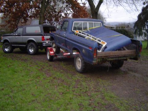 1979 Ford Bronco - A gray Ford Bronco is parked next to a blue pickup truck that is towing a trailer loaded with a blue tarped object.