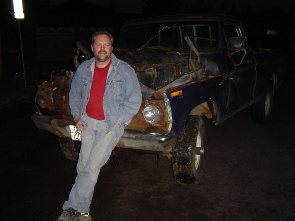 Rusty blue pickup truck with hood open, man in denim jacket leaning on front fender