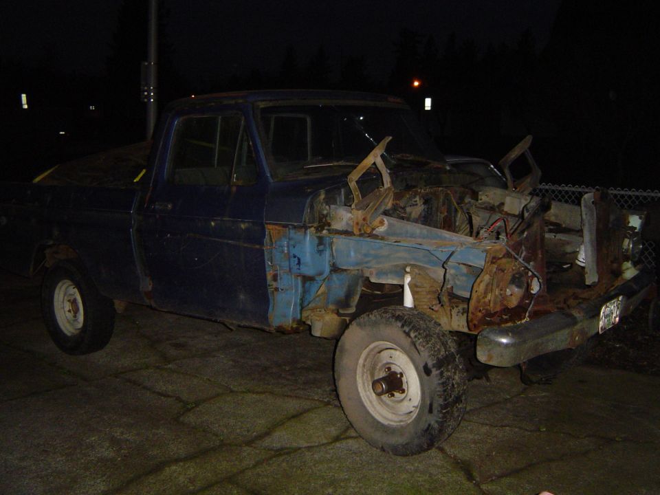 beat-up blue pickup truck missing its hood and fenders, parked at night