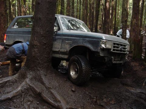 1987 Ford Bronco - A 1987 Ford Bronco is stuck in thick mud off-road, with people attempting to extract it from the forest.
