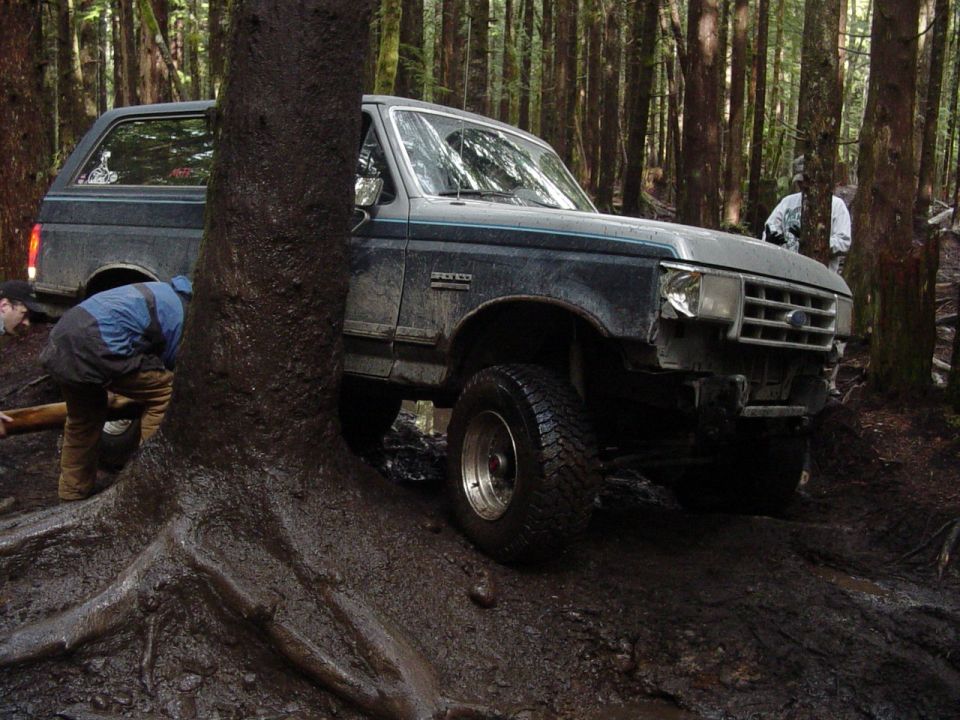 Dark blue 1987 Ford Bronco stuck in mud, front bumper missing, driver side door open, people trying to free it