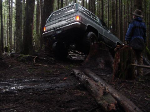 1987 Ford Bronco - A gray Chevrolet K5 Blazer navigates a muddy, wooded trail with a person watching.