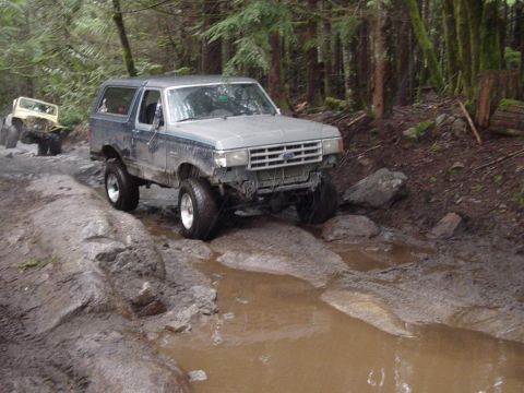 1987 Ford Bronco - Two off-road vehicles traverse a challenging, wet, and wooded path.