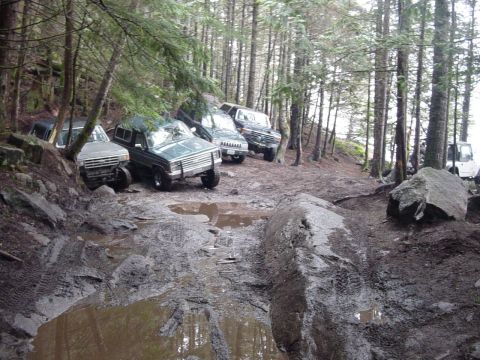 1987 Ford Bronco - Several off-road vehicles are seen on a challenging, muddy trail surrounded by trees.