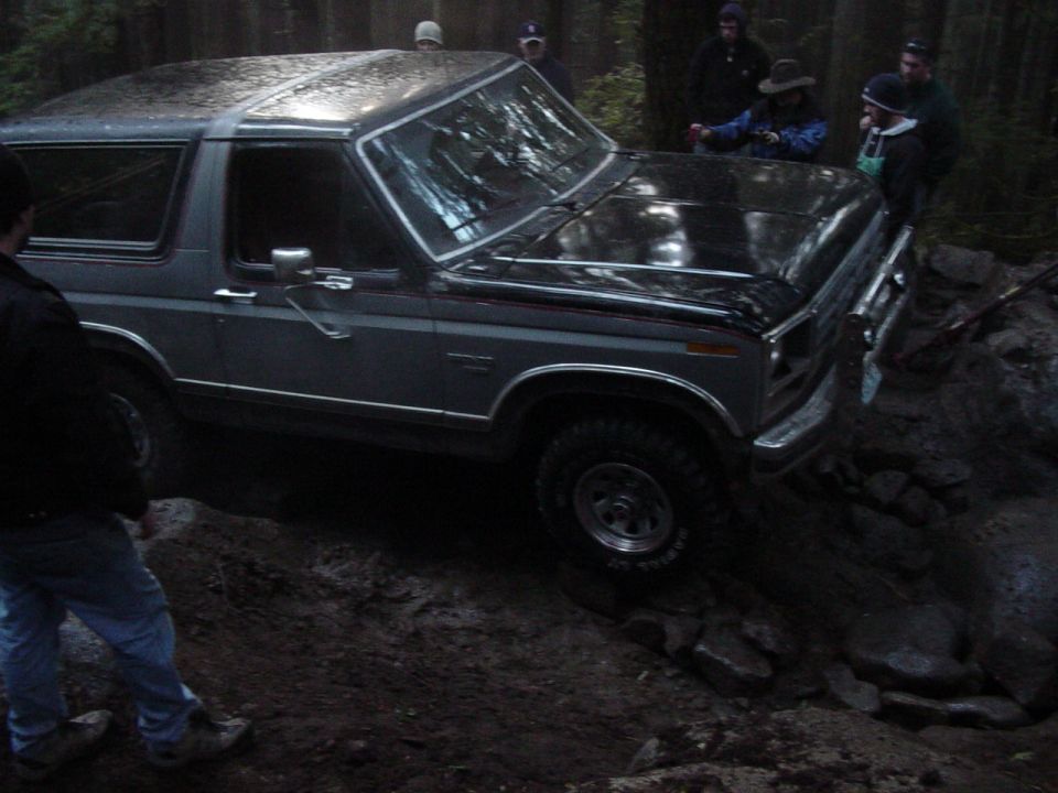 Silver and gray Ford Bronco on a rocky wooded trail with onlookers watching