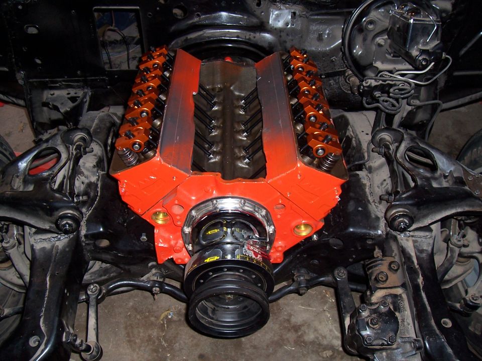 Orange small-block V8 in a stripped engine bay with valve covers off showing roller rockers