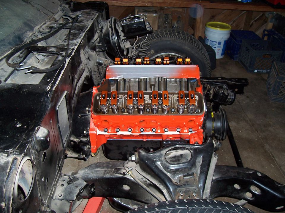 Orange small-block Chevy with valve covers off showing rockers and bare aluminum intake