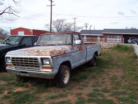 1979 Ford F-100 - A weathered 1979 Ford F-100 pickup truck sits in a yard, showing significant rust and primer gray paint.