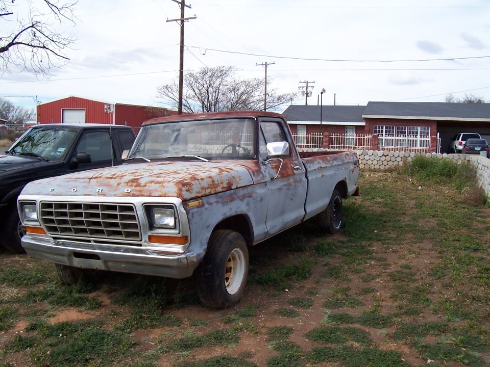 Rusty 1979 Ford F-100 pickup truck, primer gray with rust patches, parked on dirt lot next to a black truck