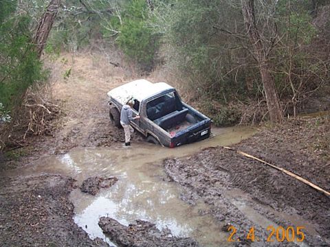 1986 Ford Bronco - A pickup truck is stuck in a muddy ditch off a wooded trail.