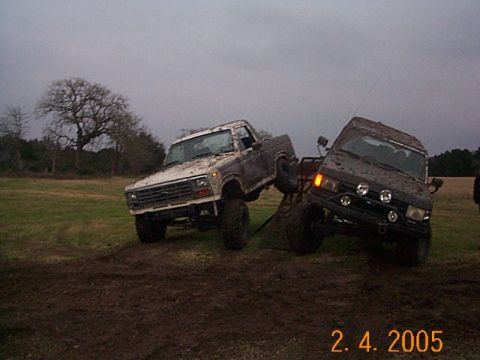 1986 Ford Bronco - Two heavily muddied off-road vehicles are stuck in a field under an overcast sky.