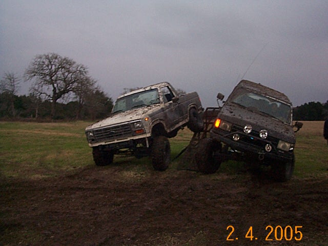 Mud-covered 1980s pickup truck angled on a mound of dirt next to a dark SUV