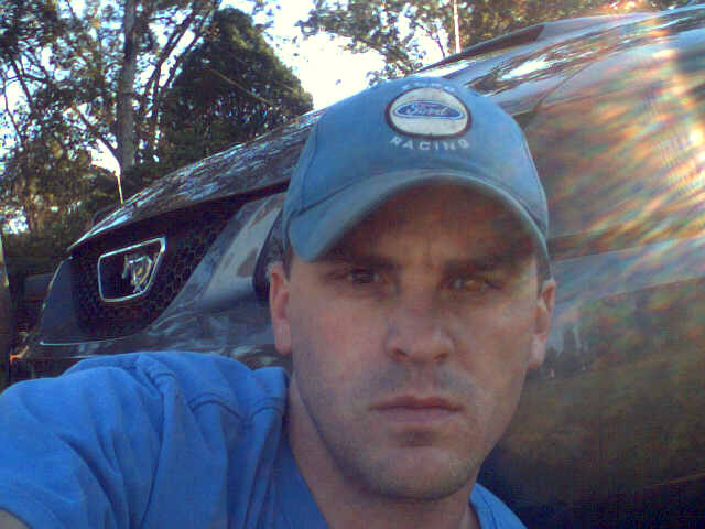 Man in blue shirt and Ford Racing cap in front of a dark colored car with a honeycomb grille and a horse emblem
