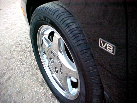 Close-up of a car's front fender showing a V8 badge and a chrome wheel with a tire. The car is dark colored