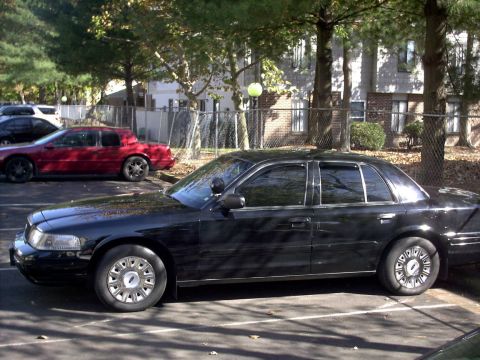 2001 Ford Crown Victoria - A black 2001 Ford Crown Victoria sedan is parked in a lot next to a red coupe.