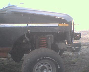 1983 Ford Bronco - Front corner view of a dark SUV, showing off-road tires and suspension components. The SuperMotors logo is visible in the lower right.