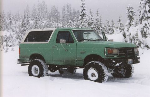 1990 Ford Bronco - A green Ford Bronco with a white top is parked in a snowy forest.