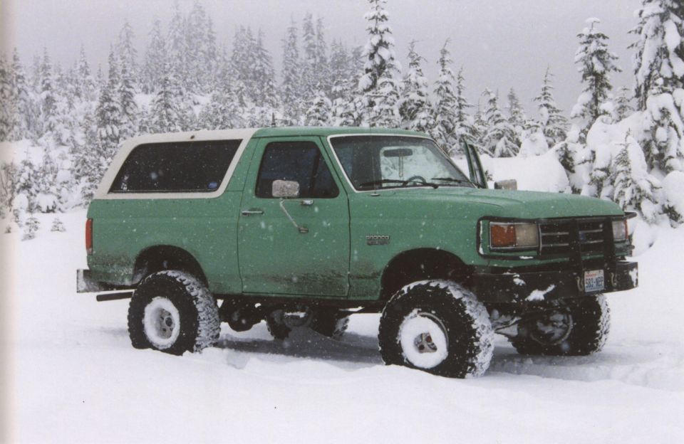 Green lifted Ford Bronco with white top parked in deep snow among snowy evergreens