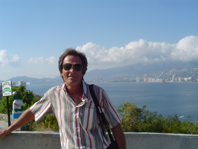 Man in sunglasses and striped shirt posing at a scenic overlook above a coastal bay with city skyline behind.