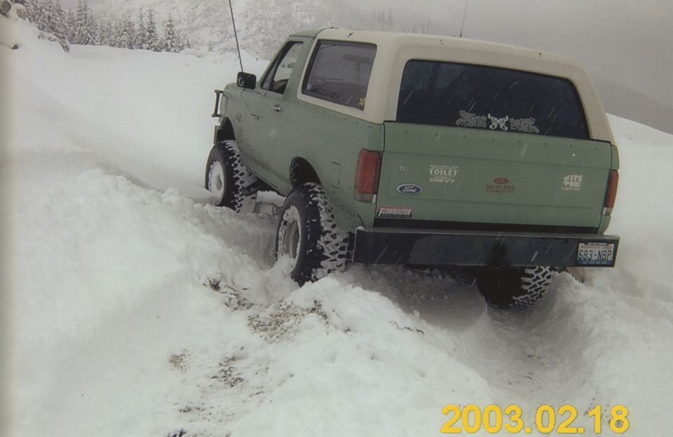 Green Ford Bronco with white hardtop stuck axle-deep in snow, rear 3/4 view