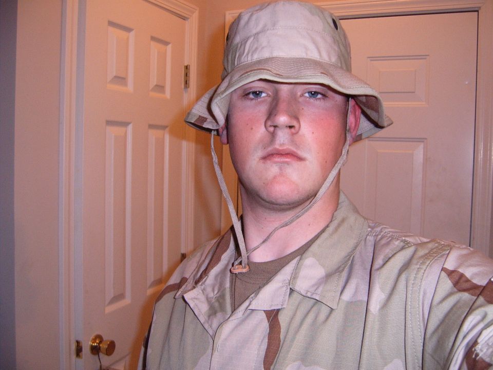 man in desert camouflage uniform and boonie hat, standing indoors in front of a white door