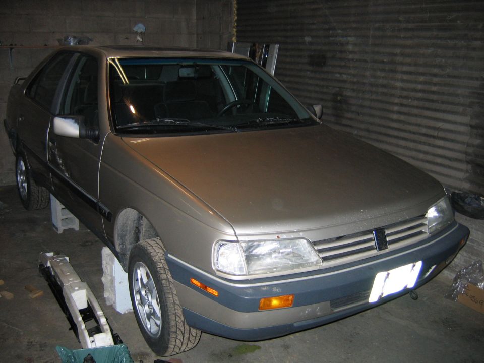 Gold sedan parked front-3/4 in a garage, gray lower trim, parts on floor