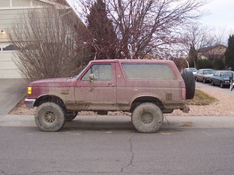 1991 Ford Bronco - A lifted, mud-covered 1991 Ford Bronco is parked on the street in front of a house.
