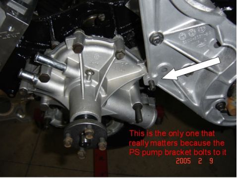 1990 Ford Bronco - Close-up of a cast aluminum water pump and front accessory bracket bolted to the front of a black engine block, with a white arrow pointing…