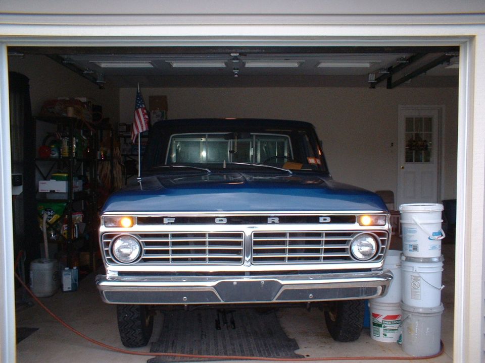 Blue 1970s Ford pickup front view in a garage, chrome grille with FORD hood letters and American flag on the antenna