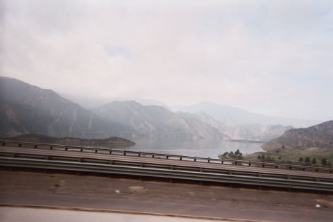 1990 Ford Bronco - hazy mountain reservoir and cloud-covered ridges seen across a metal highway guardrail from a moving vehicle.