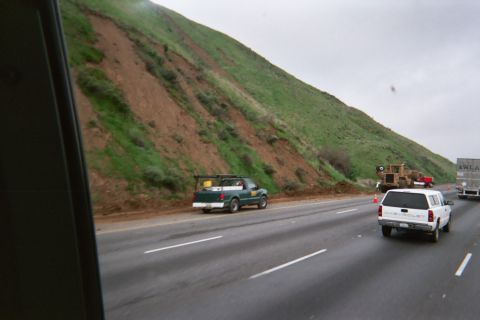 1990 Ford Bronco - green pickup and a white pickup travel a freeway below a steep eroded green hillside, seen from inside a vehicle.