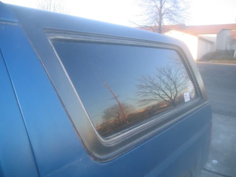 1981 Ford Bronco - Side view of a blue pickup truck's camper shell window, showing reflections.