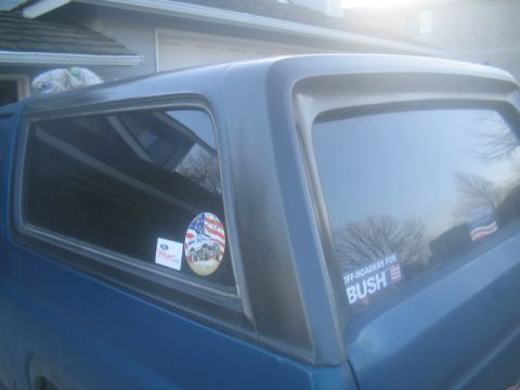 1981 Ford Bronco - A blue pickup truck with a camper shell is parked in front of a garage. Stickers adorn the rear window.