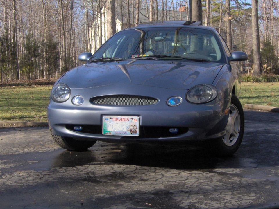 Front view of a blue-gray Ford Taurus sedan with Virginia Jamestown plate on a driveway among bare trees