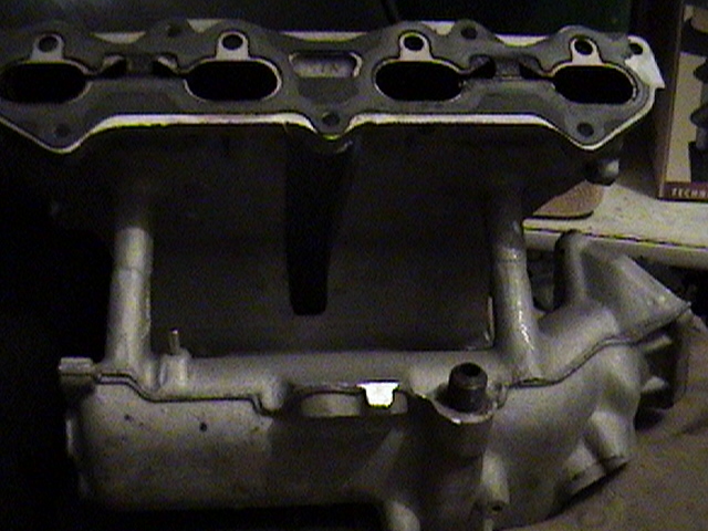 Bare aluminum four-cylinder intake manifold on a bench, port face toward camera