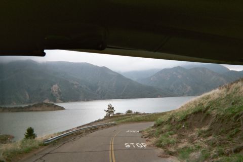1990 Ford Bronco - curving hillside road marked STOP overlooks a misty reservoir and fog-draped mountains, viewed from inside a vehicle.