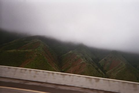 1990 Ford Bronco - Steep green-and-rust colored hills disappearing into low fog, seen above a concrete highway barrier from a vehicle.