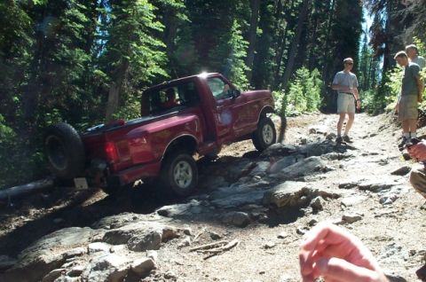 1992 Ford F-150 - A red 1992 Ford F-150 navigates a challenging, rocky off-road trail surrounded by trees.