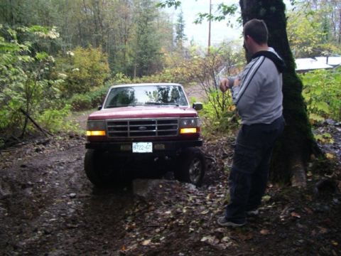1992 Ford F-150 - A red 1992 Ford F-150 navigates a rough, wooded path, with a man standing by a tree observing.