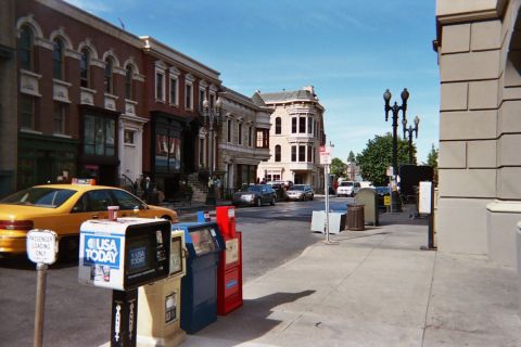 1990 Ford Bronco - yellow taxi and USA Today newspaper boxes line a sunny studio-backlot street with ornate building facades.