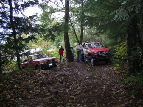 1992 Ford F-150 - Two off-road vehicles, a red Ford F-150 and a red SUV, are parked on a muddy trail in a wooded area.