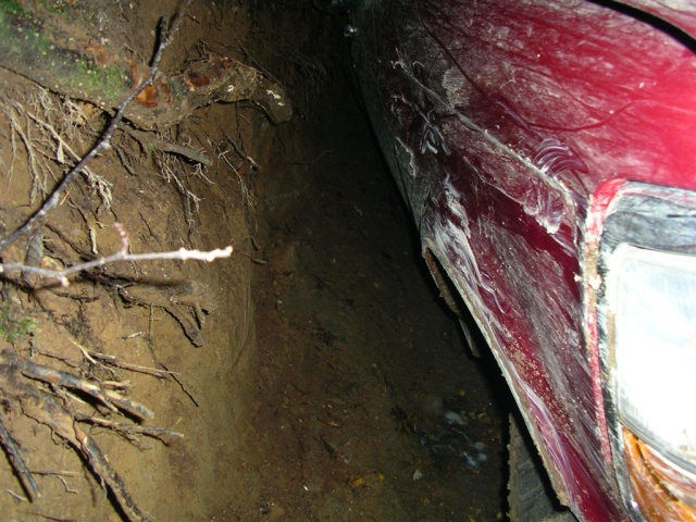 Red vehicle fender and headlight assembly partially buried in dirt with tree roots visible