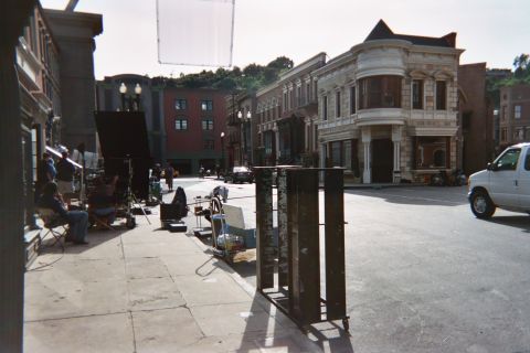1990 Ford Bronco - Crew members and lighting equipment occupy a sidewalk on a studio-backlot street corner beside a white van.