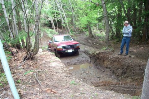 1992 Ford F-150 - A red pickup truck is shown driving through a muddy rut in a wooded area. A person stands on the bank watching.