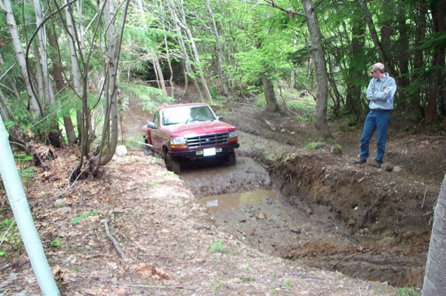 Red pickup truck navigating a muddy ditch on a wooded trail. Guy watches