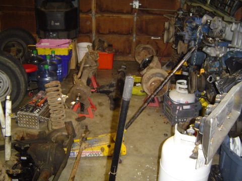 1989 Ford Bronco - A cluttered garage displays various vehicle parts, including axles, springs, and an engine, suggesting a repair or restoration project.