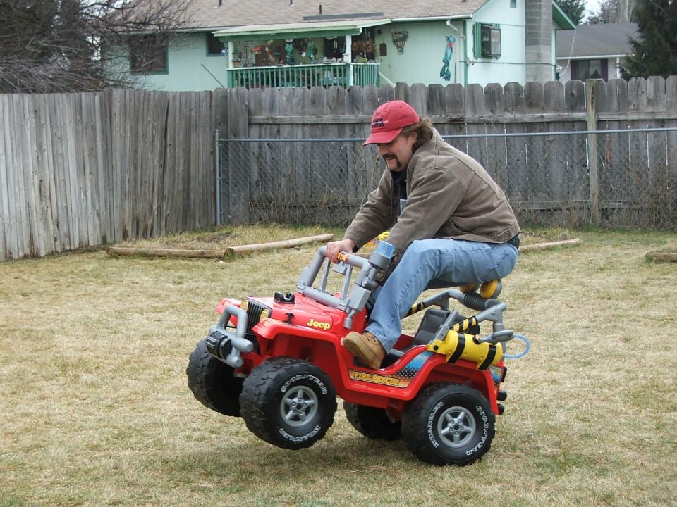 Man riding a red Jeep child's ride-on toy across a backyard lawn