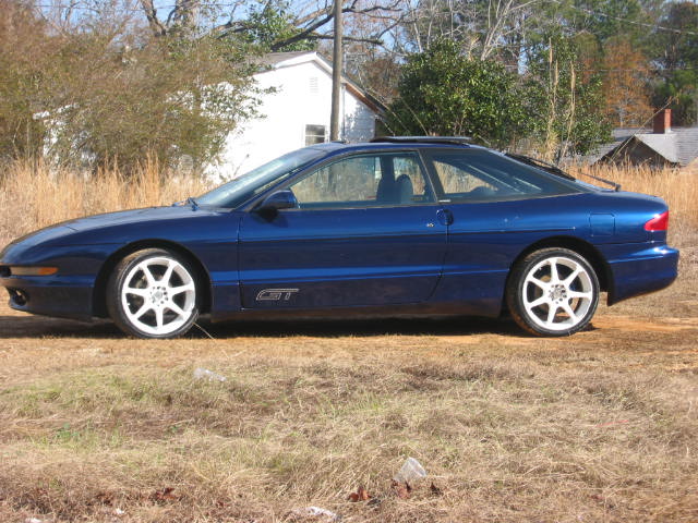 Blue coupe in side profile with GT badge and white alloy wheels parked on grass.