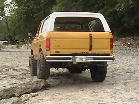1986 Ford Bronco - Rear view of a yellow Ford Bronco with a white top, driving on a rocky path.