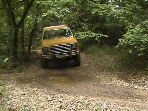 1986 Ford Bronco - A yellow van navigates a muddy dirt track surrounded by dense foliage.